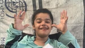 Save Rimas in Gaza – Rimas smiling and raising her hands while sitting in front of a tent.