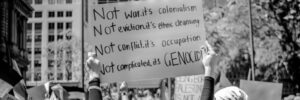 Protesters holding signs at a pro-Palestine rally, with one sign reading “Not war, it’s colonialism. Not eviction, it’s ethnic cleansing. Not conflict, it’s occupation. Not complicated, it’s genocide.”