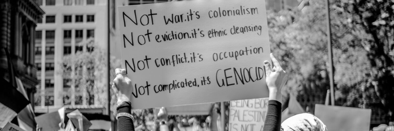 Protesters holding signs at a pro-Palestine rally, with one sign reading “Not war, it’s colonialism. Not eviction, it’s ethnic cleansing. Not conflict, it’s occupation. Not complicated, it’s genocide.”