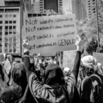 Protesters holding signs at a pro-Palestine rally, with one sign reading “Not war, it’s colonialism. Not eviction, it’s ethnic cleansing. Not conflict, it’s occupation. Not complicated, it’s genocide.”
