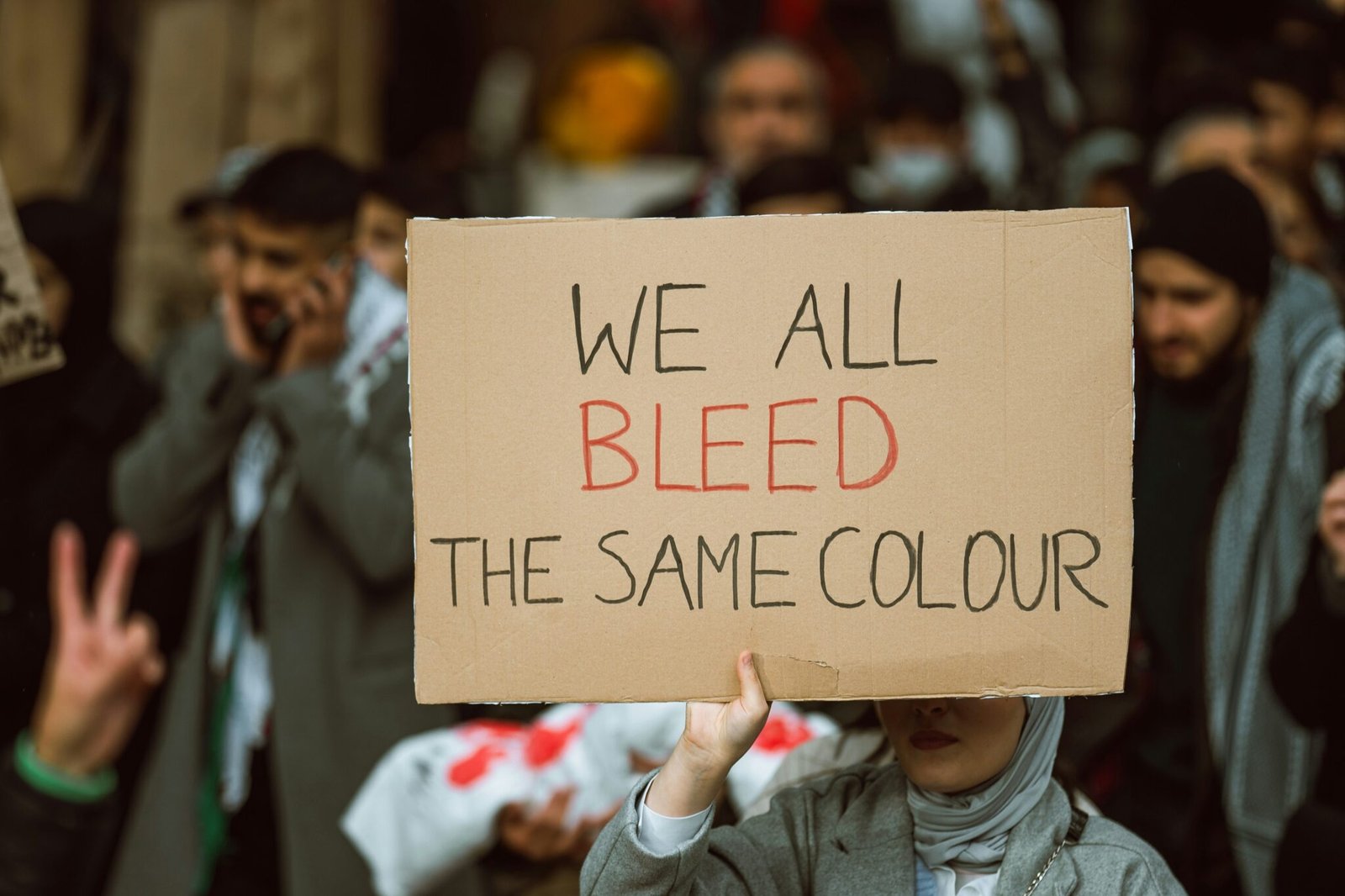 A woman in a light grey hijab holds a cardboard sign reading “WE ALL BLEED THE SAME COLOUR,” with “BLEED” written in red, amid a blurred crowd of protesters.